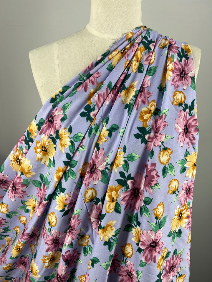 A versatile choice for any occasion, the Super Cheap Fabrics' Printed Rayon - Watercolour 2.0 - 145cm is draped over a mannequin. The light purple background is adorned with yellow and pink flowers and green leaves, elegantly arranged in folds over the mannequin's shoulder.