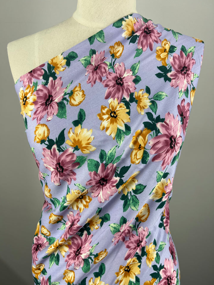 A mannequin adorned in a one-shoulder garment made from Super Cheap Fabrics' Printed Rayon - Watercolour 2.0 - 145cm, featuring a lively and colorful design with large pink, yellow, and green leaves on a light purple background. This vibrant floral pattern stands out beautifully against the neutral background, making it a versatile choice.