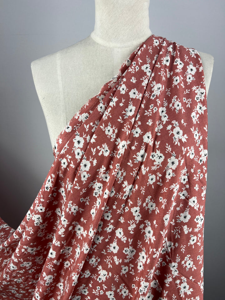 Close-up of a fabric draped over a mannequin torso. The Printed Rayon - Mona Lisa by Super Cheap Fabrics is light red with a white floral pattern, showcasing small flowers and leaves evenly distributed across the material. This versatile, multi-use textile stands out against the plain, neutral grey background.