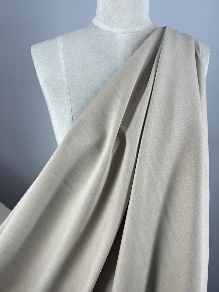 The Designer Suiting - Sandy from Super Cheap Fabrics, expertly draped over a pale grey dress form, showcases the smooth and flowing texture of this medium to heavy weight suiting material against a plain backdrop.