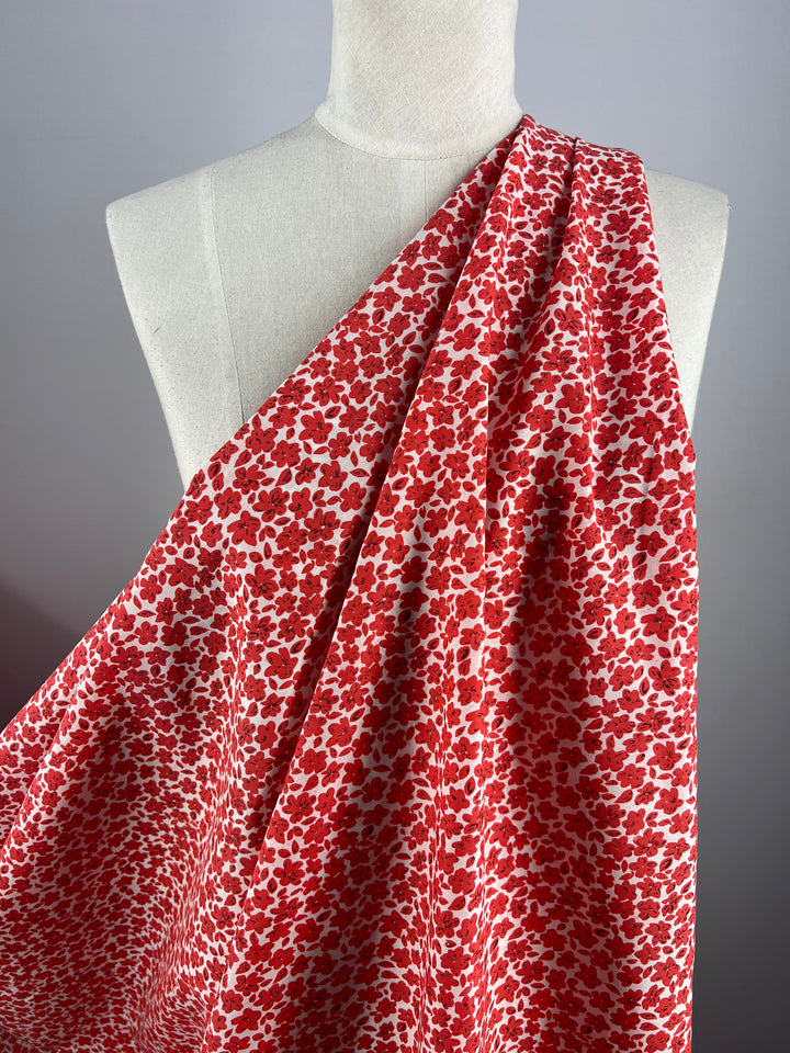 A mannequin draped with Super Cheap Fabrics' Printed Cotton - Red Rain - 145cm, adorned with a dense, all-over pattern of small white flowers. The lightweight fabric is displayed over one shoulder, cascading downward to showcase its texture and vivid red hue against a neutral gray background.