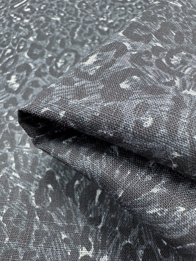 A close-up of a folded piece of "Linen Blend - Cool Jungle - 140cm" by Super Cheap Fabrics showcases its dark gray, lightweight material with a marbled, textured pattern of lighter gray and white streaks. This breathable fabric is displayed on a matching background, offering a seamless, layered look ideal for summer outfits.