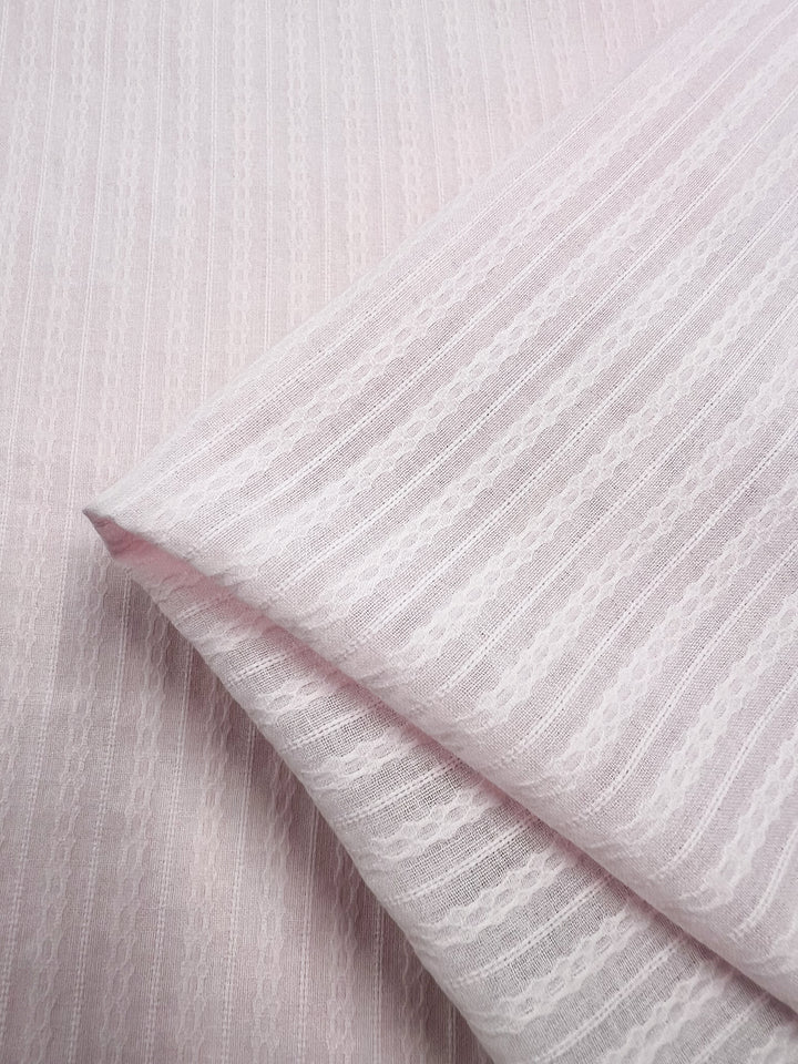 A close-up view of a folded piece of Textured Cotton - Shrinking Violet - 140cm from Super Cheap Fabrics shows the light pink, striped 100% cotton fabric. This extra light weight cotton features intricate, lace-like vertical stripes alternating with sheer bands, creating a delicate and elegant texture.