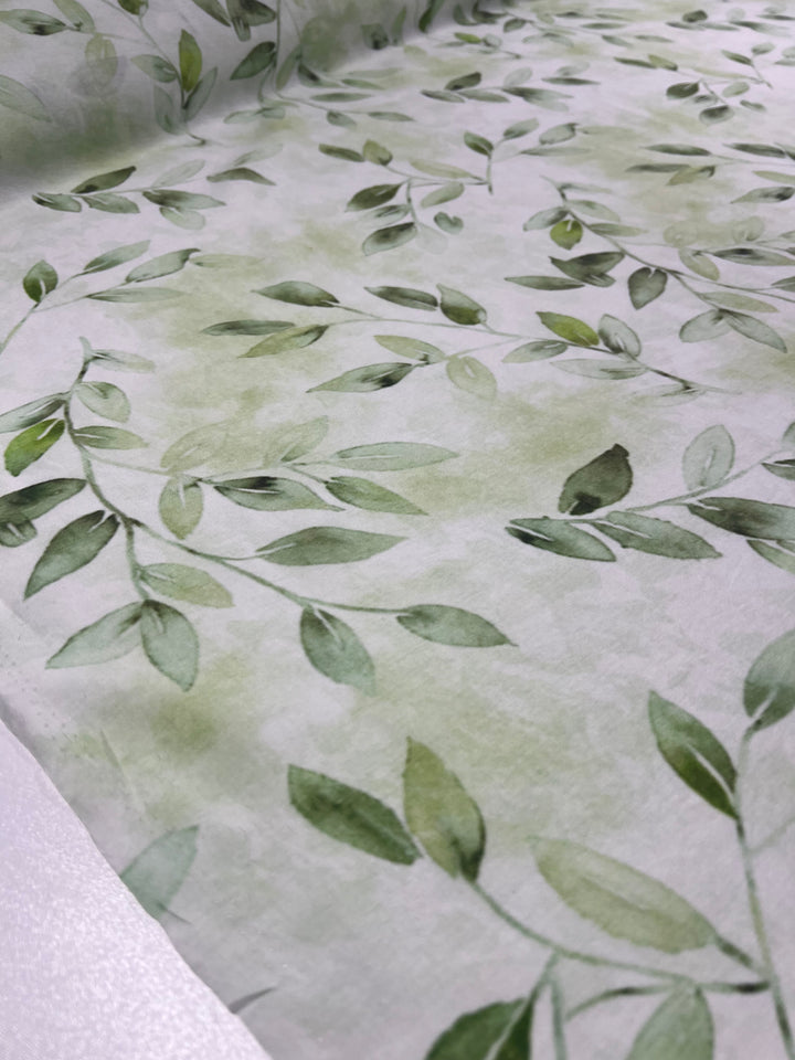 A close-up view of Super Cheap Fabrics' Designer Cotton - EUCAS - 150cm reveals a lightweight cotton fabric adorned with a botanical print. The design showcases delicate green leaves and vines on a light, almost watercolor-like background, creating a serene and nature-inspired pattern perfect for luxury sewing projects.