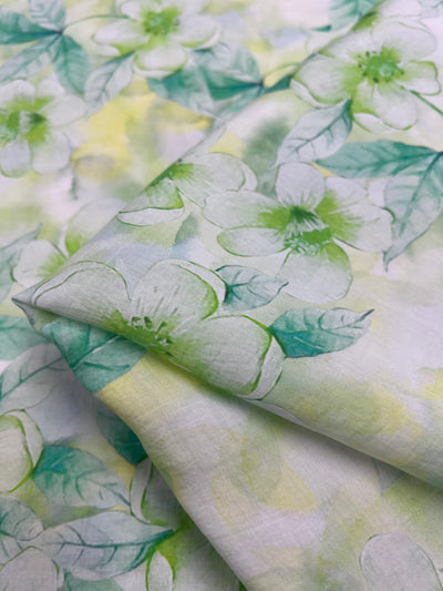 A close-up view of Super Cheap Fabrics' Designer Cotton - Periwinkle, featuring large white flowers with green centers and leaves on a soft watercolor-style background in shades of green and yellow. Ideal for luxury sewing projects, this breathable fabric is neatly folded to display its multiple layers.