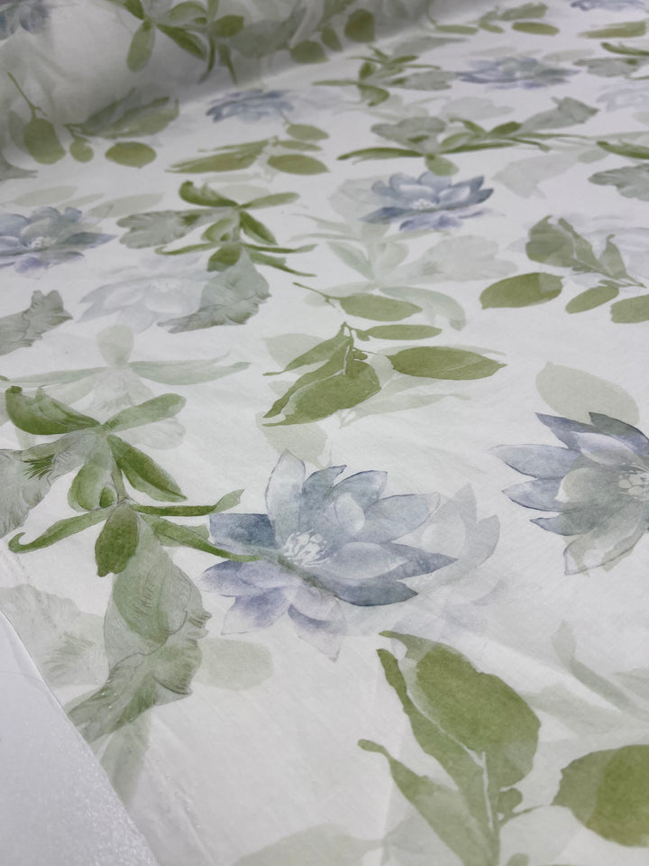 A close-up shot of Super Cheap Fabrics' Designer Cotton - Lotus - 150cm. Perfect for luxury sewing projects, this fabric features pale blue flowers with green leaves and stems set against a white background. The design is soft and watercolor-like in appearance, making it an ideal choice from designer cotton fabrics.