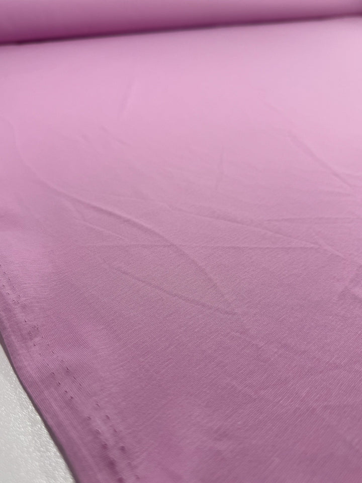 A close-up of Super Cheap Fabrics' Cotton Jersey - Pink Lavender (155cm) shows its lightweight, smooth surface with subtle creases, lying flat on a white background.