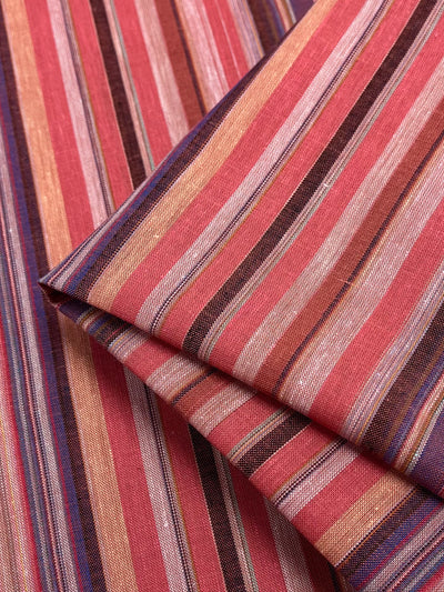 Close-up of the folded Linen Blend - Colombo - 150cm fabric from Super Cheap Fabrics, showcasing a vibrant striped pattern in shades of red, pink, orange, purple, and brown. This yarn-dyed design features stripes of varying widths for a visually dynamic effect. The slightly textured finish imparts a vintage look to this woven fabric.