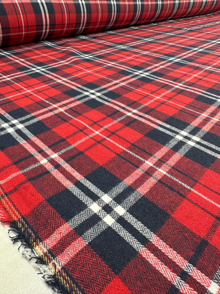 A roll of Super Cheap Fabrics' Suiting - Trad Tartan - 147cm fabric with a traditional tartan pattern in black, white, and red is partially unrolled on a table. The smooth and neatly laid out surface showcases the symmetrical checkered design, making it an ideal choice for formal suits.