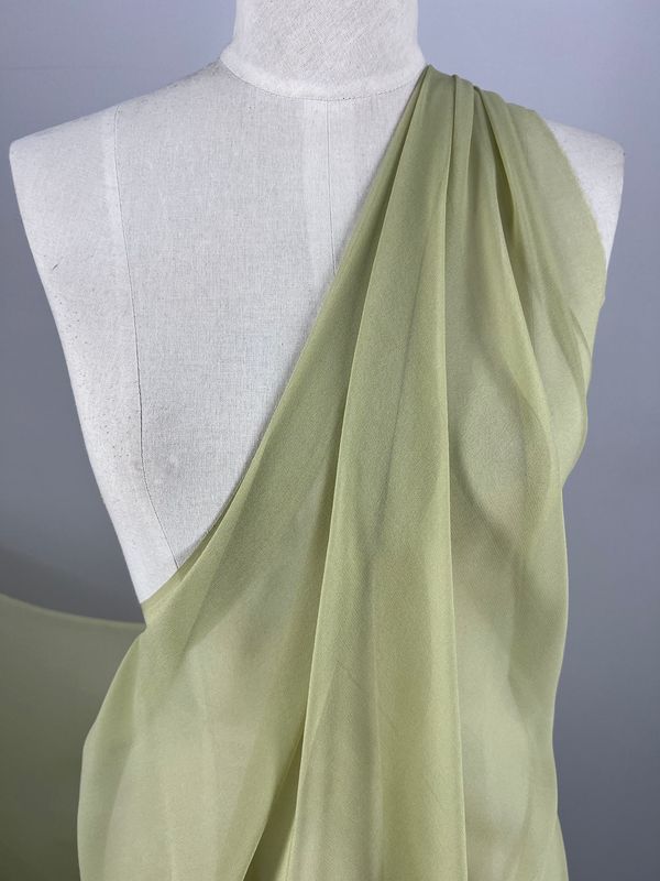 A dress form mannequin draped with lightweight, celery green sheer fabric arranged asymmetrically over its shoulder, creating a flowing, elegant effect. Perfect for spring and summer outfits, the Silk Georgette - Celery from Super Cheap Fabrics adds a touch of sophisticated charm against the neutral background.