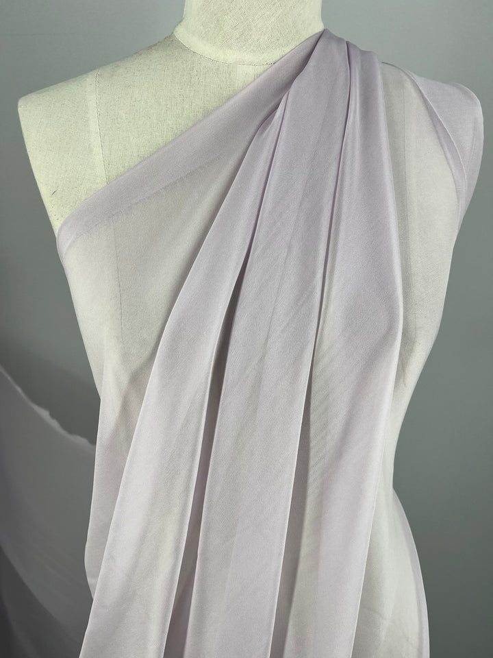 The Orkid Silk Georgette Fabric from Super Cheap Fabrics, with a width of 135cm, draped elegantly over a white dress form in a toga-like style featuring soft, flowing folds. The lightweight, sheer lavender fabric creates a delicate and graceful summer outfit against a plain, neutral background.