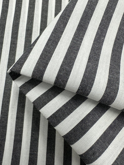 Close-up of black and white striped fabric with a folded section on top of a flat section. The lightweight fabric displays alternating vertical stripes, creating a classic and bold pattern. The 100% cotton texture of Super Cheap Fabrics' Cotton Lawn - Black Stripe - 150cm appears smooth with a slightly silky finish.