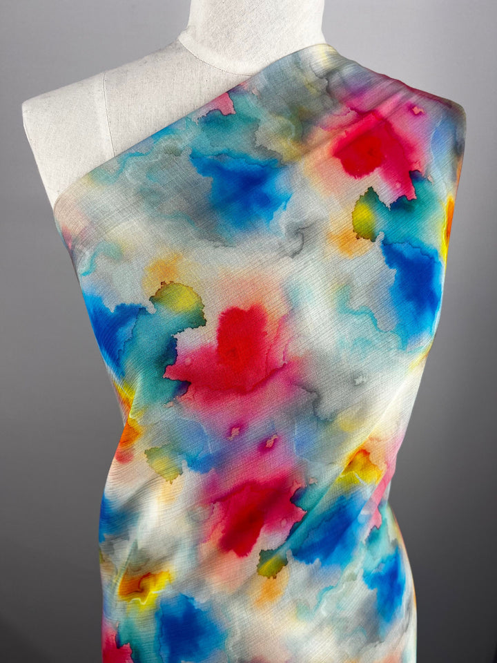 A dress form displays the Designer Cotton - Venus - 150cm fabric from Super Cheap Fabrics, featuring lively abstract watercolor patterns in shades of blue, pink, yellow, and green against a gray background—ideal for luxury sewing projects.