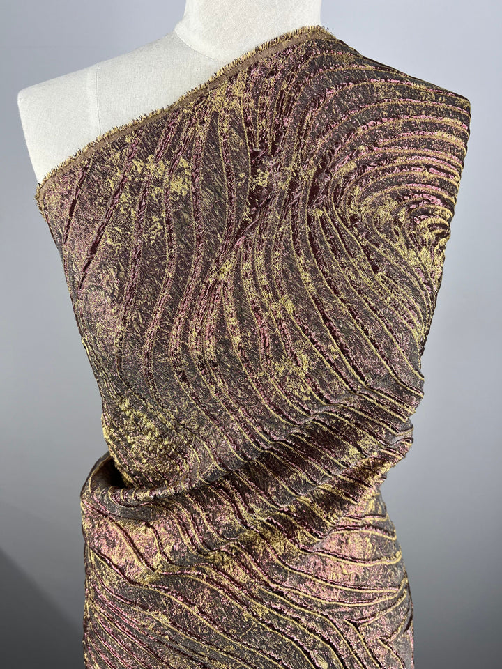 An elegant dress form is adorned with the Designer Metallic Brocade - Amelia - 150cm from Super Cheap Fabrics, showcasing a textured fabric with a swirling pattern in metallic shades of gold, bronze, and green. Crafted from lightweight brocade fabric, the design features a unique layered ripple effect that adds depth and movement reminiscent of Turkey's rich textile tradition.