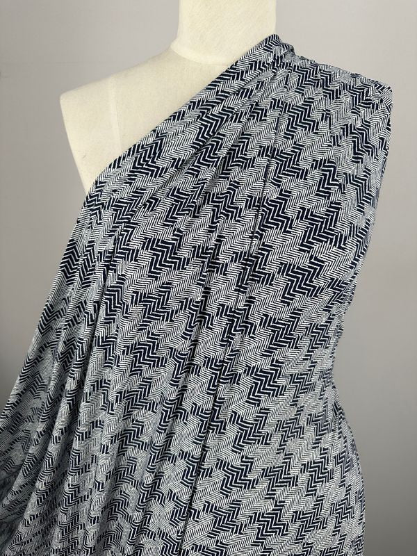 A posed mannequin draped in Super Cheap Fabrics' Printed Lycra - Tazo, featuring a medium-weight fabric with a black zigzag pattern, creates a textured, abstract design. This Polyester/Spandex blend elegantly highlights the flow and drape. The plain and neutral background emphasizes the intricate pattern of this 150cm wide fabric.