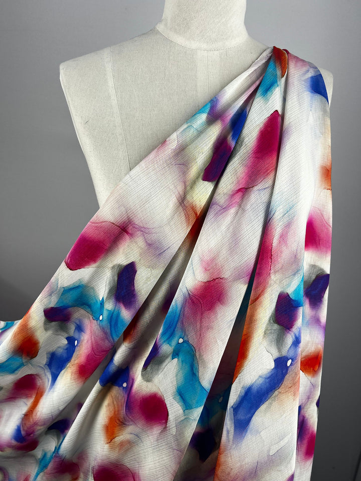 A mannequin adorned with "Designer Cotton - Crysta - 150cm" from Super Cheap Fabrics, showcasing a luxurious watercolor-patterned fabric. This material boasts bold splashes of pink, blue, purple, and orange tones on a light background, creating a fluid and artistic design ideal for luxury sewing projects that require artistic flair.