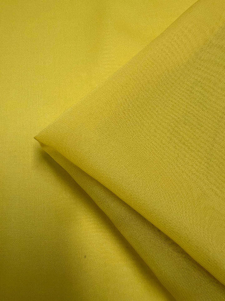 A close-up image of a piece of bright yellow, lightweight Silk Georgette - Quail - 135cm by Super Cheap Fabrics, featuring soft folds. The texture of the 100% silk georgette is slightly rough, with subtle, fine lines running throughout. The lighting highlights the vibrant color and the details of the material's surface—perfect for summer outfits.