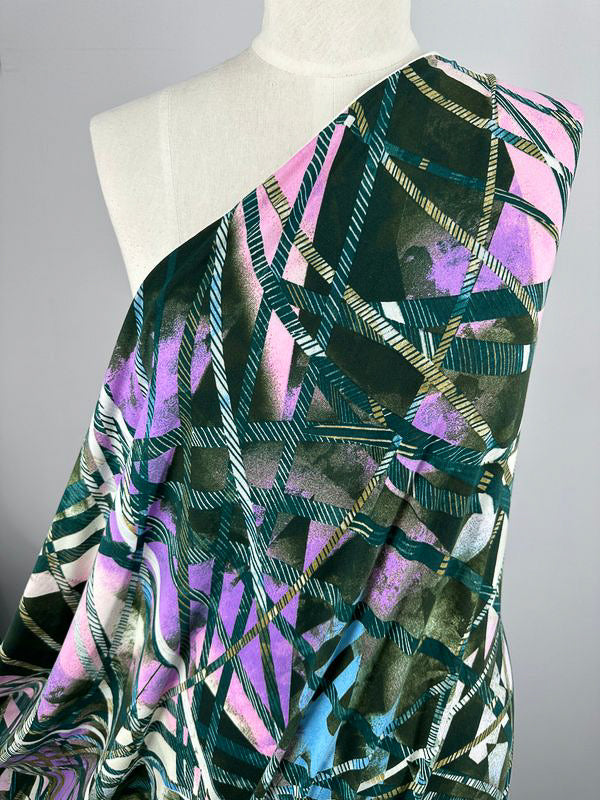 A dress form draped with Super Cheap Fabrics' Printed Lycra - Tied Up, a medium-weight fabric showcasing an abstract geometric pattern in shades of green, purple, pink, and white. The asymmetrical draping creates a one-shoulder effect, highlighting the dynamic crisscrossing lines and vibrant colors.