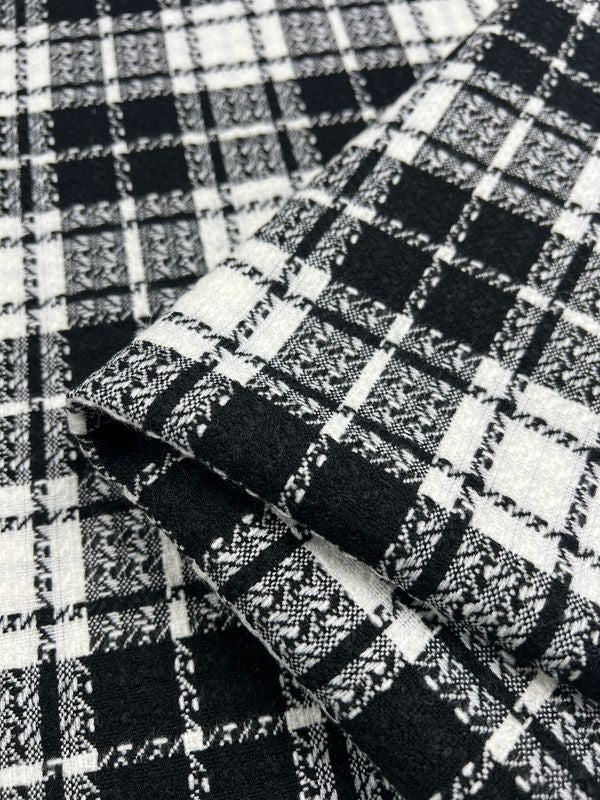 Close-up of the Designer Tweed - Chanel fabric from Super Cheap Fabrics, showcasing a black tartan pattern with intersecting woven designs in varying shades and textures. The picture focuses on a folded section, highlighting the detail and intricacy of this heavy weight 100% polyester textile, ensuring durability and strength. This fabric measures 145cm wide.