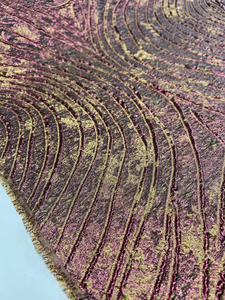 A close-up of a textured surface featuring a wavy, organic pattern in shades of pink, gold, and brown. The design consists of raised lines and metallic elements reminiscent of light weight brocade fabric, creating a visually intricate and tactile feel. The background appears smooth and light blue. This description perfectly matches the Designer Metallic Brocade - Amelia - 150cm from Super Cheap Fabrics.