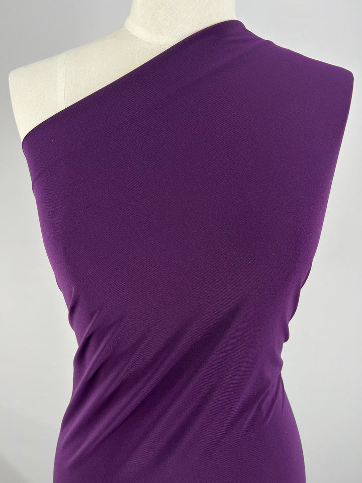 A close-up of a mannequin wearing a deep purple, one-shoulder dress crafted from Super Cheap Fabrics' ITY Knit - Plum - 150cm. The medium weight fabric appears smooth and slightly stretchy, draping elegantly to highlight the form. The background is plain and light-colored, ensuring the purple dress stands out prominently.