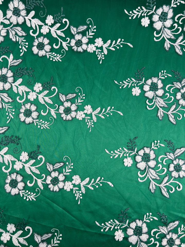 The "Embroidered Lace - Teal - 150cm" from Super Cheap Fabrics is a teal, lightweight material adorned with white floral patterns and leafy vines. This detailed embroidery features various flower types, forming a delicate and intricate lace-like design that spans the entire fabric.