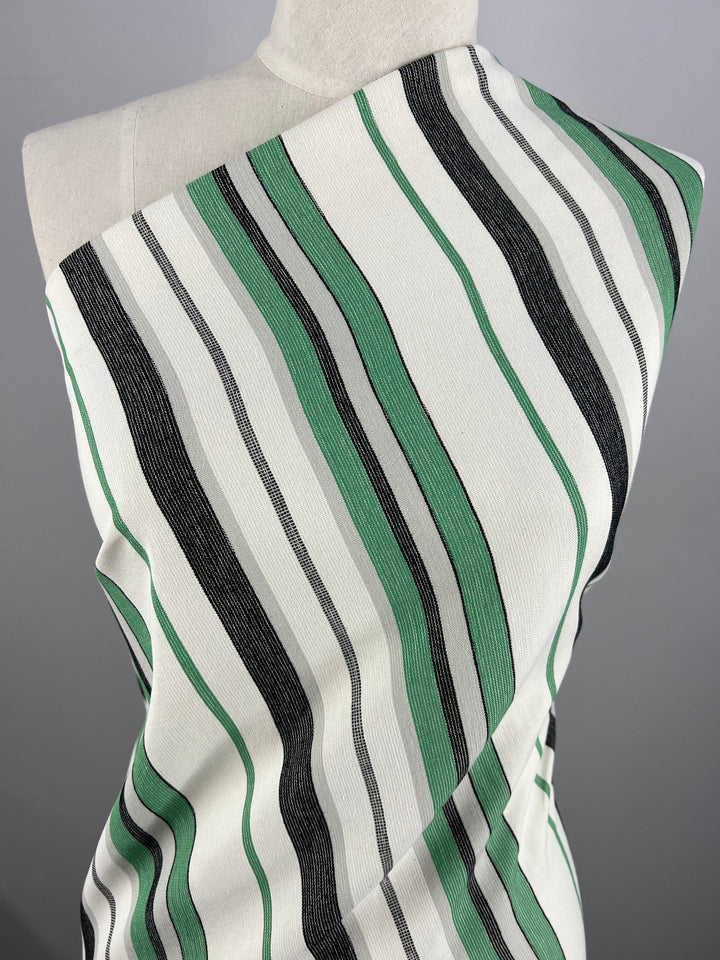 A mannequin is draped with a fabric from the Super Cheap Fabrics Designer Cotton - Green Rock Candy - 145cm, featuring a pattern of wavy vertical stripes in green, black, and white. The stripes vary in width and create an undulating, flowing appearance across the fabric. The background is plain and grey.