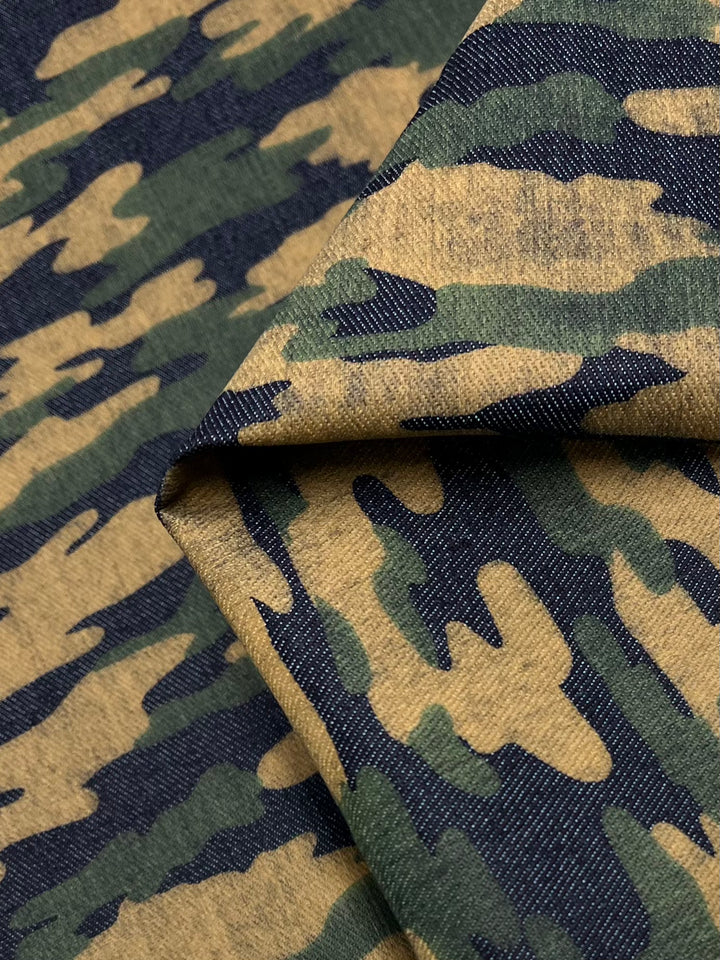 A close-up of a folded piece of medium weight Printed Denim - Camouflage from Super Cheap Fabrics. The 145cm wide cotton fabric features abstract shapes in shades of navy green, brown, and tan.
