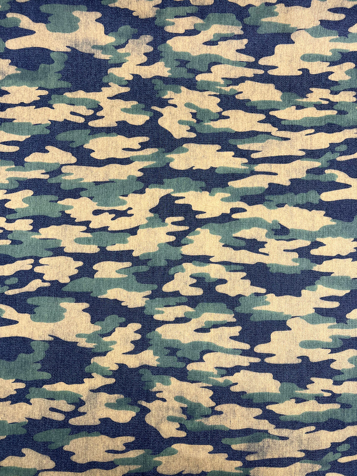 A detailed close-up of the "Printed Denim - Camouflage - 145cm" fabric by Super Cheap Fabrics, showcasing a blend of dark green, light green, navy green, brown, and beige splotches in medium weight. The irregular shapes are artfully combined to mimic a natural outdoor environment for optimal concealment.