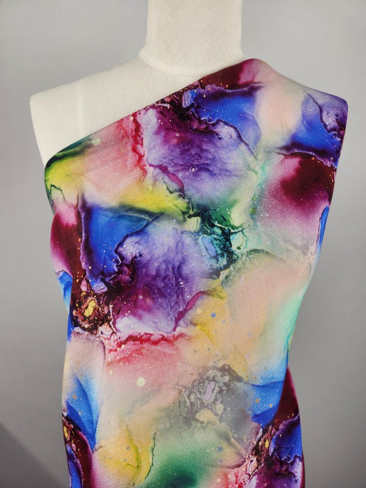 A mannequin adorned with the Designer Cotton - Saphira - 150cm from Super Cheap Fabrics showcases a vibrant, abstract-patterned textile featuring splashes of purple, blue, yellow, pink, and green. The colorful design resembles swirling watercolors, offering an artistic and dynamic appearance ideal for luxury sewing projects.