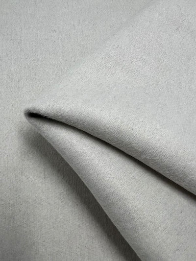 Close-up of Super Cheap Fabrics' Wool Cashmere - Speckley Snow, 150cm, folded at an angle. The light gray material appears smooth with a soft texture, showcasing subtle variations in shading and light.