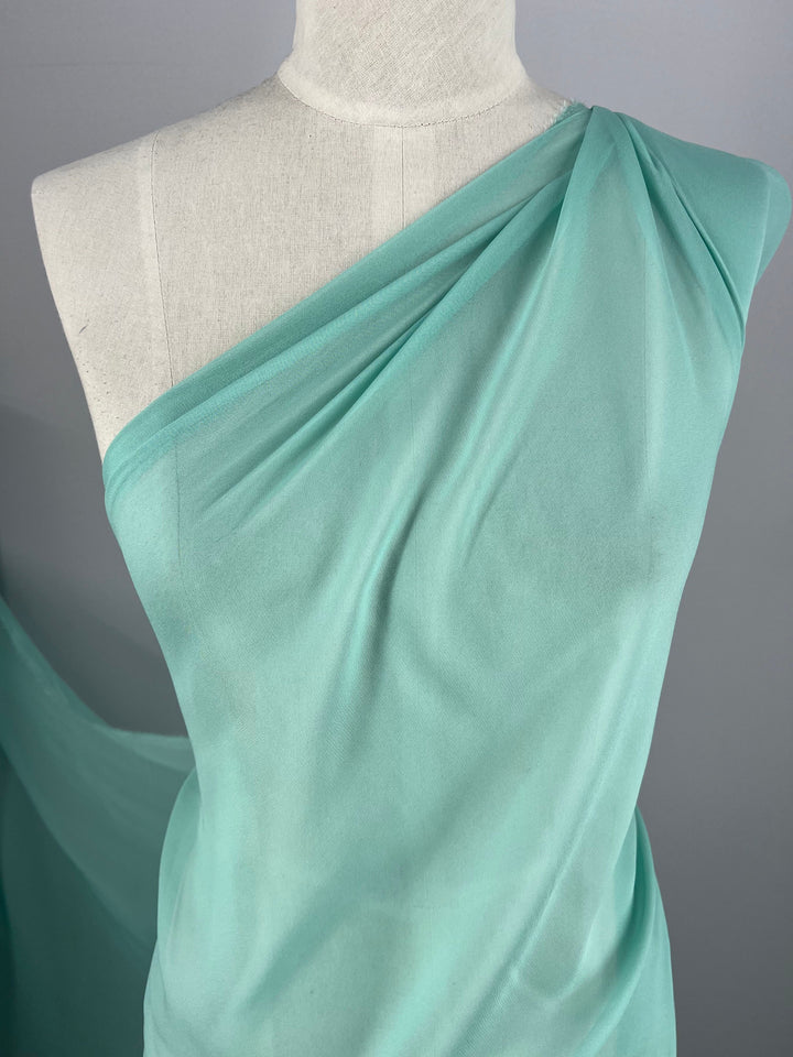 Close-up of a mannequin draped with Super Cheap Fabrics' Silk Georgette - Lagoon - 135cm over one shoulder, forming a Grecian-style look. The light green, semi-transparent fabric has gentle folds and a soft, flowing appearance. Perfect for spring and summer outfits, the background is plain and neutral.