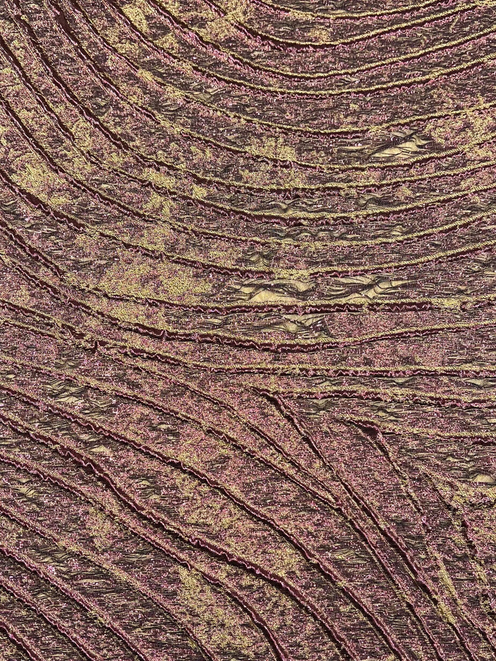 A close-up of an abstract texture featuring shimmering gold and reddish-pink hues. The surface has a wavy, ridged pattern that resembles flowing lines or topographical maps, similar to the intricate details found in Super Cheap Fabrics' Designer Metallic Brocade - Ameila - 150cm. The colors blend and vary in intensity, giving a dynamic and vibrant metallic visual effect.