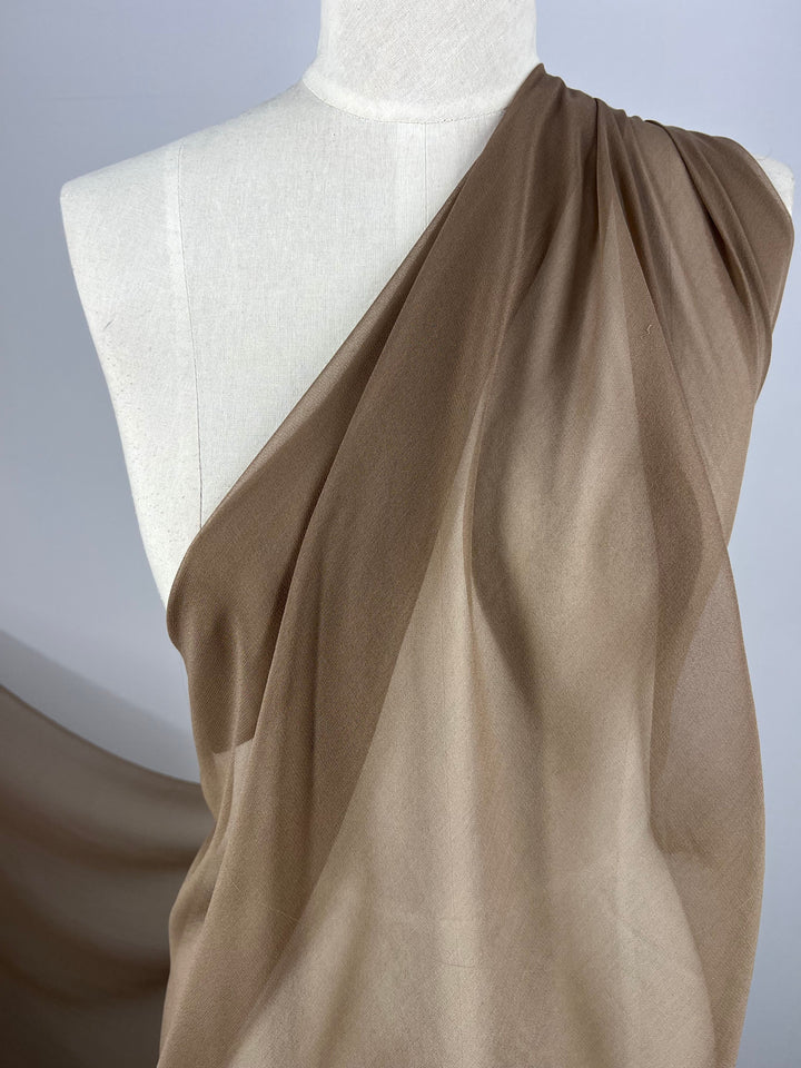 A white mannequin is adorned in a sheer, delicate Silk Georgette - Bark fabric from Super Cheap Fabrics. The lightweight material elegantly cascades over one shoulder and across the torso, forming gentle folds and a flowing look. Set against a plain gray background, the texture and drape are accentuated, making it ideal for spring and summer ensembles.