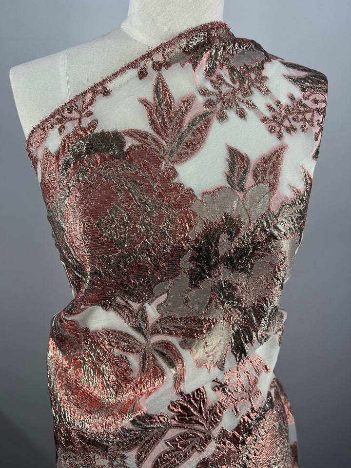 The mannequin is adorned with the Designer Metallic Brocade - Anne, a lightweight fabric from Super Cheap Fabrics. This selection features intricately patterned, large metallic floral designs in shades of copper and silver. The shiny fabric is displayed against a plain grey background, highlighting its luxurious finish.
