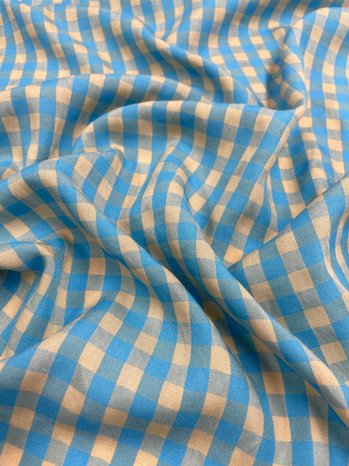 A close-up of the lightweight Cotton Poly - Cream & Blue Gingham fabric from Super Cheap Fabrics draped in soft folds. The pattern displays a grid of alternating cream and blue squares, creating a classic gingham design ideal for pants and skirts. The texture appears smooth, with slight shadowing highlighting the fabric's contours.