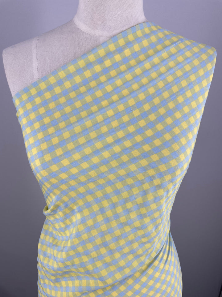 A mannequin is draped in an airy blue and yellow gingham pattern fabric from the Super Cheap Fabrics collection. The Cotton Poly - Blue & Yellow Gingham - 150cm fabric is wrapped asymmetrically around the mannequin, exposing one shoulder. The background is a plain, light gray.