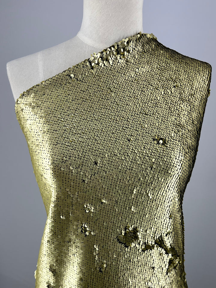 A dress form is draped with an asymmetric, one-shoulder garment made from Super Cheap Fabrics' Premium Sequins in Matte Gold - 150cm. The heavy weight fabric's texture catches the light, creating a glimmering effect. Some sequins appear loose or missing, adding a distressed look to the material.