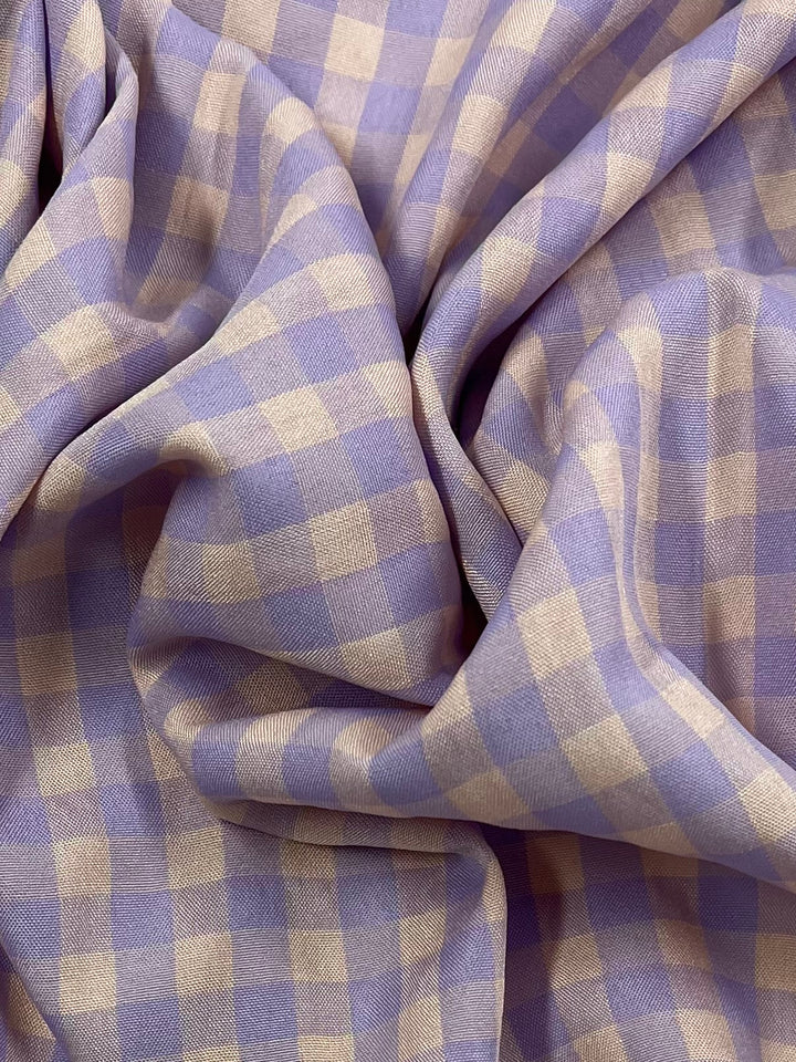 A close-up view of gathered fabric featuring a checkered pattern in pastel lavender and peach whip colors. The Cotton Poly - Peach & Purple Gingham - 150cm by Super Cheap Fabrics appears soft and slightly crumpled, creating light and shadow variations on its surface.