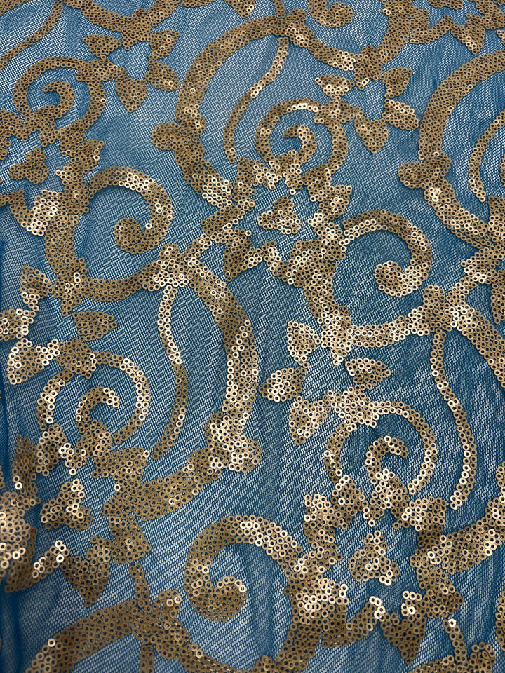 A close-up view of a fabric product, Designer Sequins- Bold Lotus - 150cm by Super Cheap Fabrics, crafted from 100% polyester and adorned with intricate golden sequin embroidery in floral and swirling patterns. The sequins form a detailed, shimmering design contrasting beautifully against the smooth blue background.