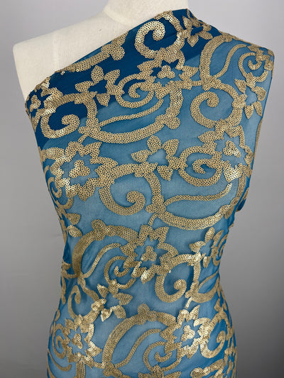 A dress form is draped with a single-shoulder, turquoise Super Cheap Fabrics Designer Sequins- Bold Lotus - 150cm featuring intricate gold sequin floral and scroll patterns. The background is plain gray.