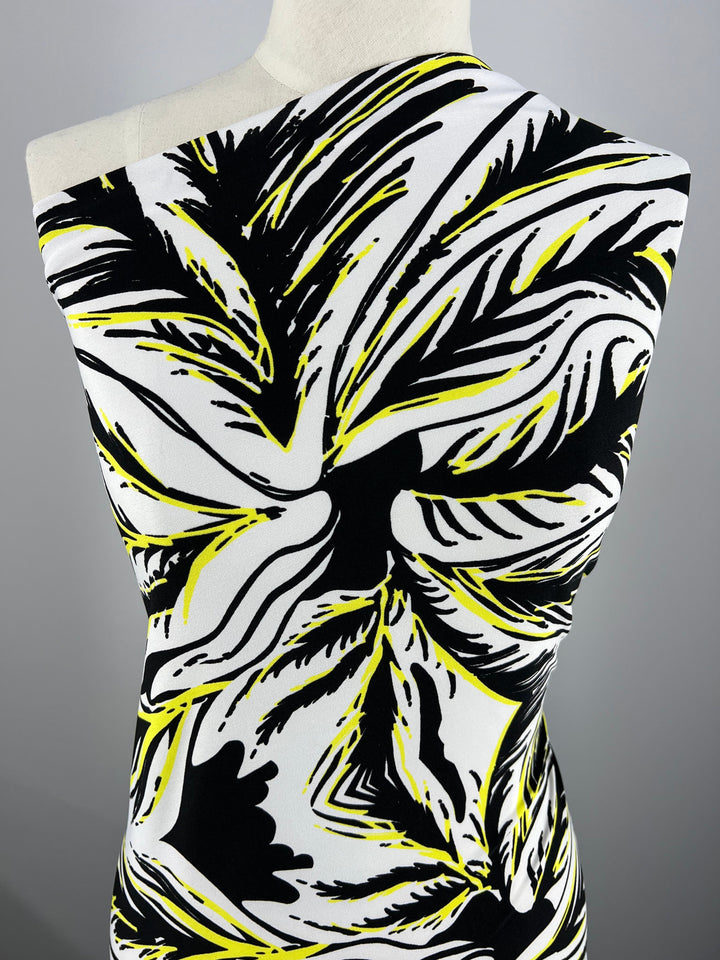 A mannequin displays a bold Printed Lycra - Yellow Palm - 150cm from Super Cheap Fabrics. The design features black, white, and yellow colors arranged in dynamic, sweeping shapes, creating a striking visual effect reminiscent of a floral formation.