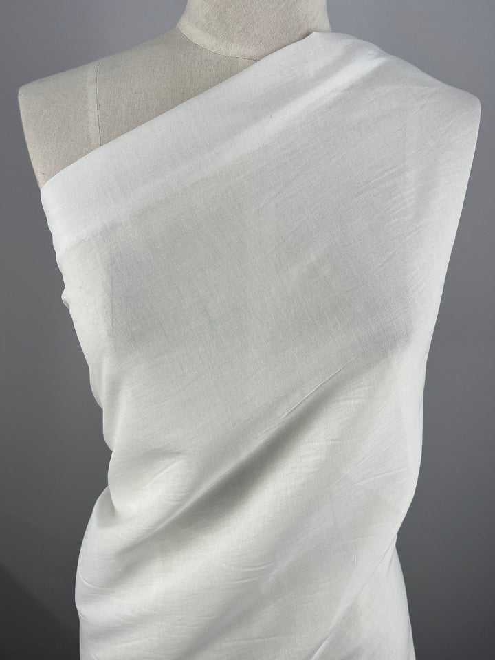 A mannequin is draped with a single piece of Super Cheap Fabrics' Cotton Voile - White - 150cm, wrapped around the body in a classic, toga-like style. The 100% Cotton fabric has a smooth texture and light sheen, creating gentle folds and a minimalist, elegant appearance against a plain grey background.